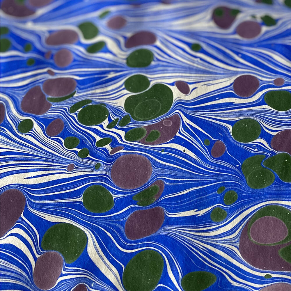 Wellenmarmorpapier /marbled paper in blau, dunkelgrün und violett. Das blau ist durchzogen mit violetten und grünen Flecken.
